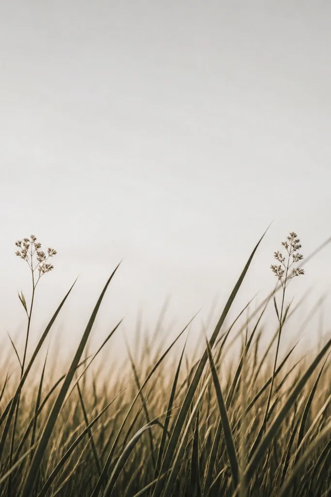 The bottom half is filled with tall, dramatic grass blades that reach upward and partially overlap each other. The top half is mostly negative space with only a couple of small flower clusters near the horizon. Grass tips are sharp and dark, with lighter blades fading upward.