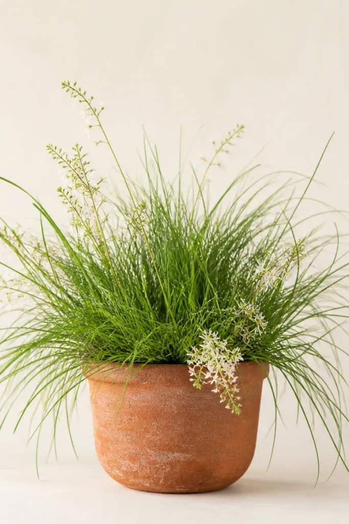 A terracotta clay pot sits at the bottom center. From inside and around the pot, grass blades spill outward. Tiny flowers sprout in a few spots, with one small cluster spilling over the pot rim. The background is pale with no extra objects.