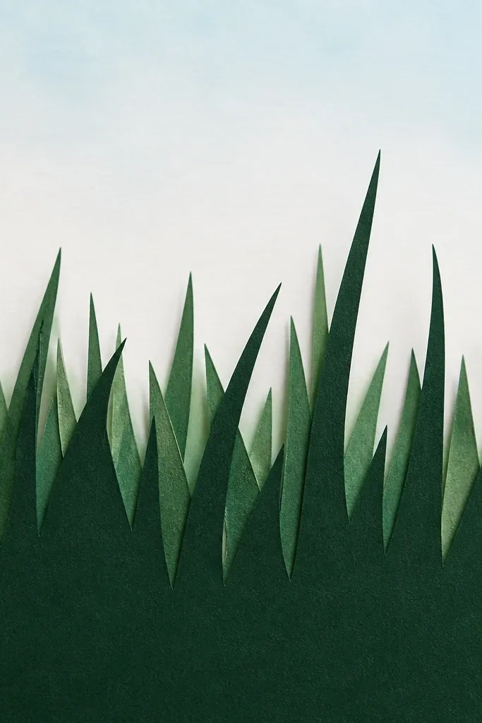 A craft piece made from cut paper. A dark grass silhouette fills the bottom half, with jagged blade edges. Behind it, a lighter backing paper creates a shadow effect. The top has a simple sky wash or blank space.