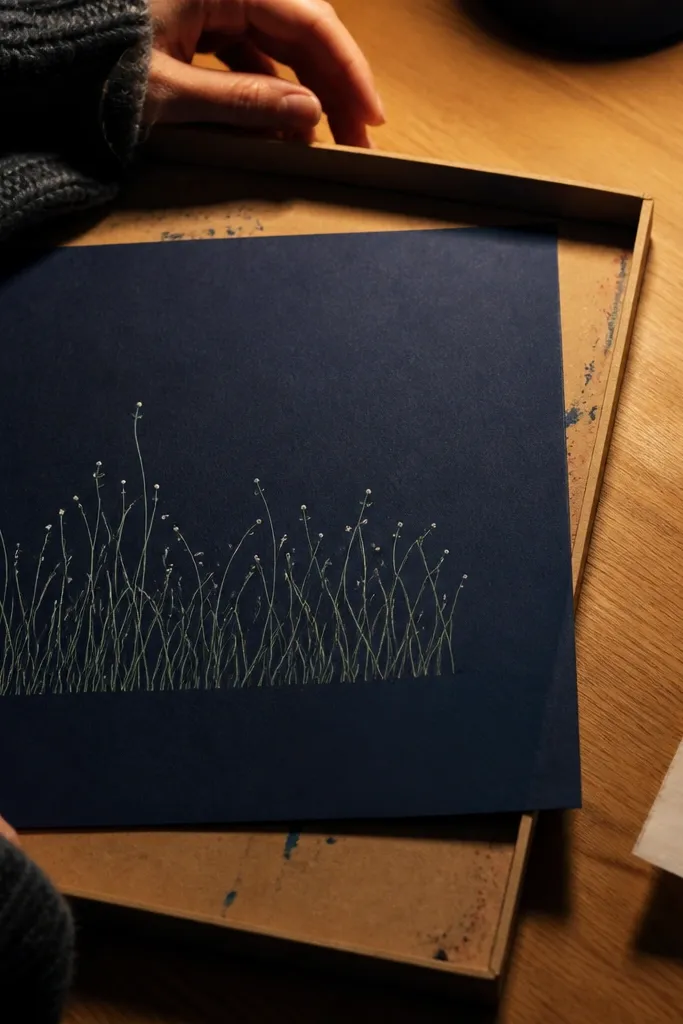 Grass blades outlined in light green on black paper, with tiny white dew dots at the blade tips. A medium dark rock sits low in the composition with a thin highlight line along its upper edge.